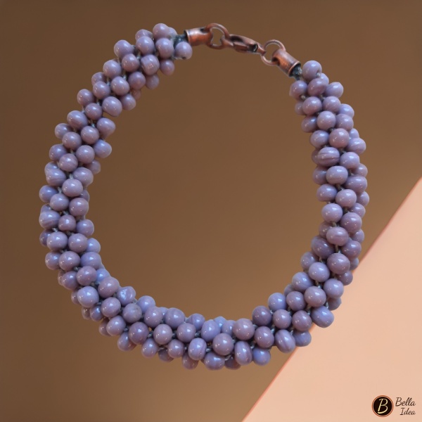 Braccialetto Kumihimo in perline color glicine di vetro lumè con chiusura ramata su sfondo rosa.